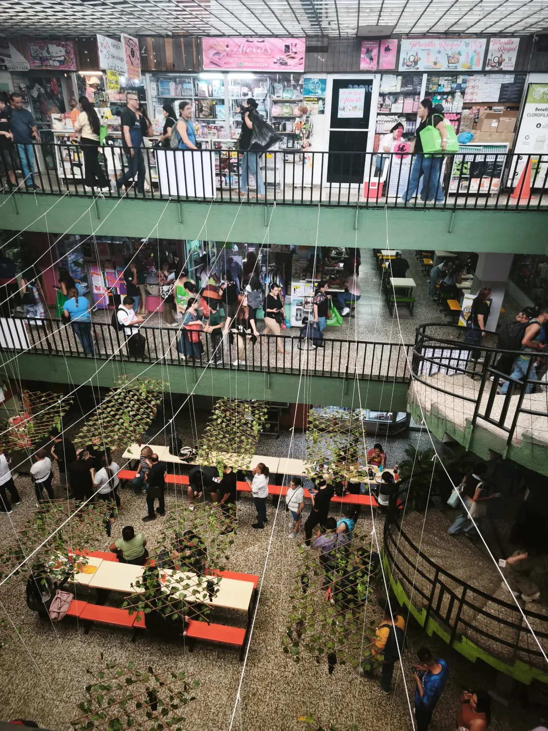 A crowded indoor market seen from above, with three floors of small shops selling cosmetics and household goods, shoppers carrying bags and food tables on the ground floor.